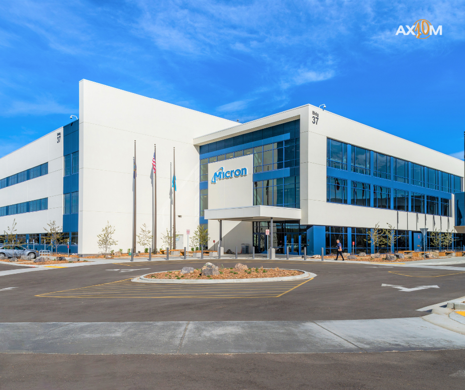 Modern office building labeled "Micron" with blue glass windows. Multiple flags outside. Two people walking. Clear blue sky above.