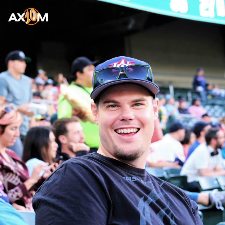 A person wearing a cap and sunglasses smiles at a sports event. Other people sit nearby. The word "AXION" is visible.