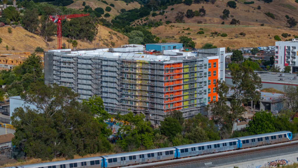 Modern modular mid-rise residential building under construction with colorful facade, scaffolding, and a red crane in the background, situated near railway tracks with a train passing by, surrounded by greenery and rolling hills.