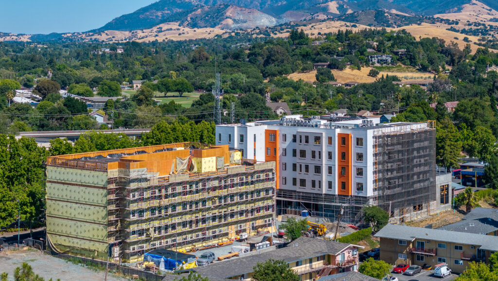 Construction site of a modular multi-story residential building with scaffolding and exterior work underway, featuring a partially completed facade with orange and white panels, surrounded by older low-rise apartments, green trees, and a mountainous landscape in the background.