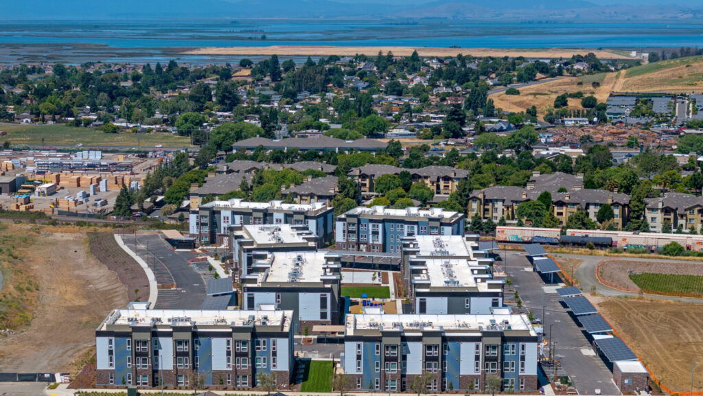 Aerial view of a modern modular multi-family residential complex with multiple three-story apartment buildings, solar panels, landscaped walkways, and a central green space, surrounded by single-family homes, construction areas, and distant water views.