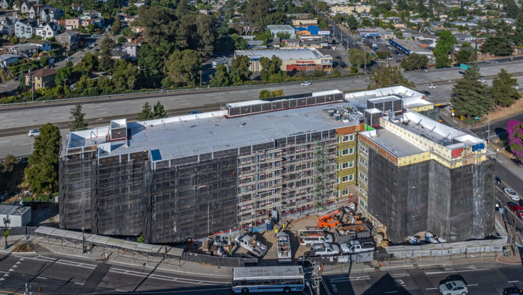 Aerial view of a large multi-story modular affordable apartment complex under construction, with scaffolding, black protective netting, and rooftop work in progress, located near a highway and surrounded by residential neighborhoods and commercial buildings.