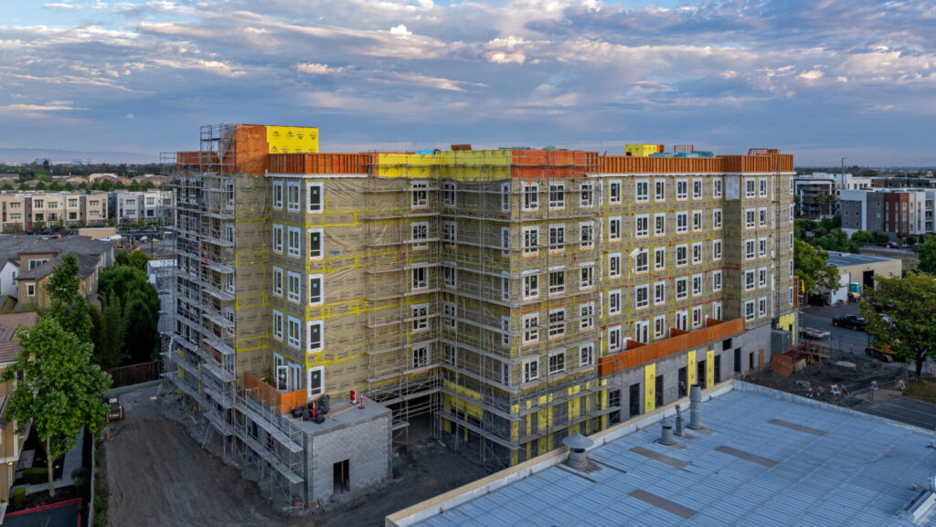 Multi-story modular residential building under construction with scaffolding, yellow weatherproof sheathing, and construction materials on-site, set against a blue sky with surrounding residential neighborhoods and commercial buildings.