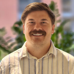 A smiling person with a mustache and striped shirt stands in front of a blurred green plant background indoors.