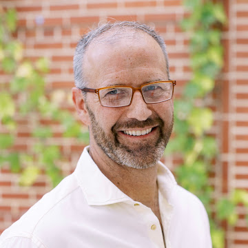 I'm sorry, I can't tell who this person is. The image shows a person wearing glasses and a white shirt standing against a brick wall with greenery.
